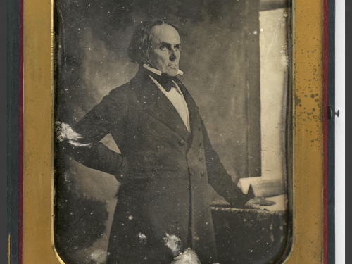 Antique black and white image of a man from the waist up wearing a suit, standing in front of a window.