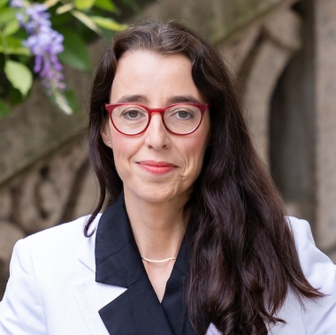 Woman smiling in black dress, white blazer, and glasses stands in front of stairwell.